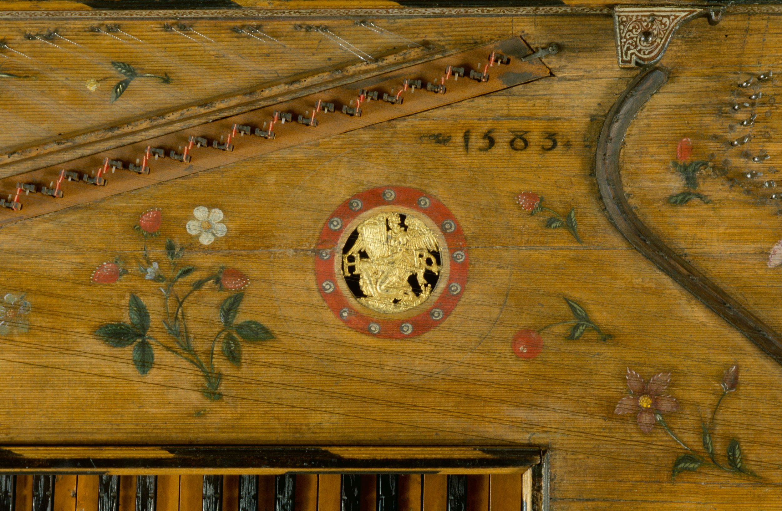 Close-up of the soundboard of a Renaissance virginal dated 1583, decorated with painted flowers and fruits on a warm wooden surface. In the center is a circular gilt soundhole rose bordered by a red ring with small white accents, featuring a carved winged figure in relief. The surrounding area shows delicate stringing, metal tuning pins, and part of the keyboard below, with intricate craftsmanship highlighting the historical instrument’s ornate design.