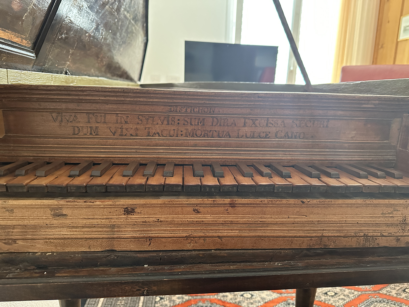 Close-up of an antique harpsichord keyboard made of wood, showing black and natural keys with visible wear. Above the keys, an engraved Latin inscription reads “Viva fui in sylvis: sum dura excussa securi: dum vixi tacui: mortua dulce cano.”