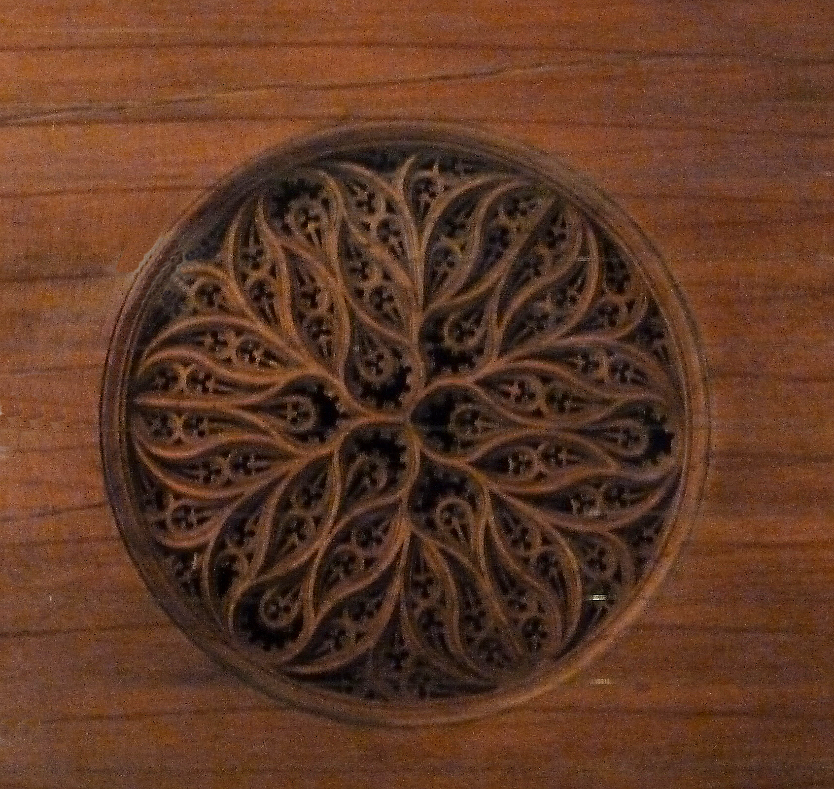 Close-up of an intricately carved wooden soundhole rose on a keyboard instrument, featuring an elaborate symmetrical pattern of swirling vines and leaf-like forms. The delicate latticework is set into a smooth wooden soundboard.