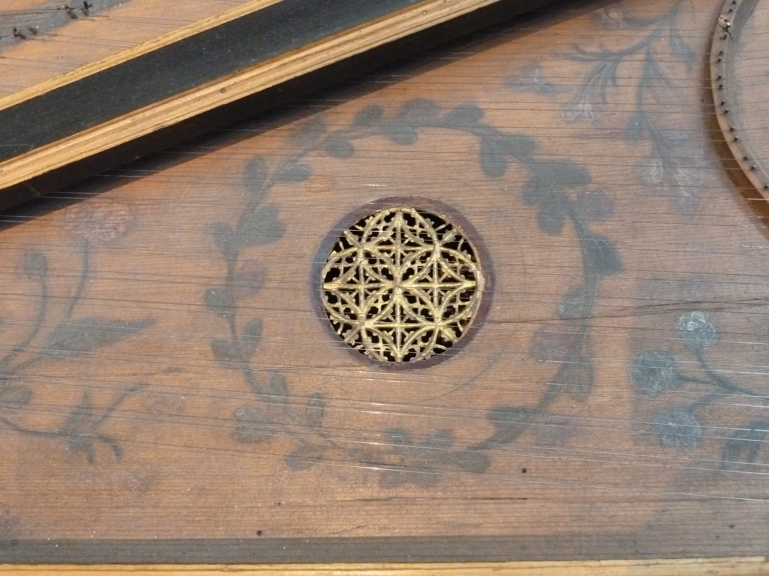 Close-up of a harpsichord soundboard showing a small circular rose soundhole with an intricate golden lattice design. Surrounding it are painted floral and leaf motifs in soft green and red tones on a warm wooden surface. Fine strings cross the image diagonally.