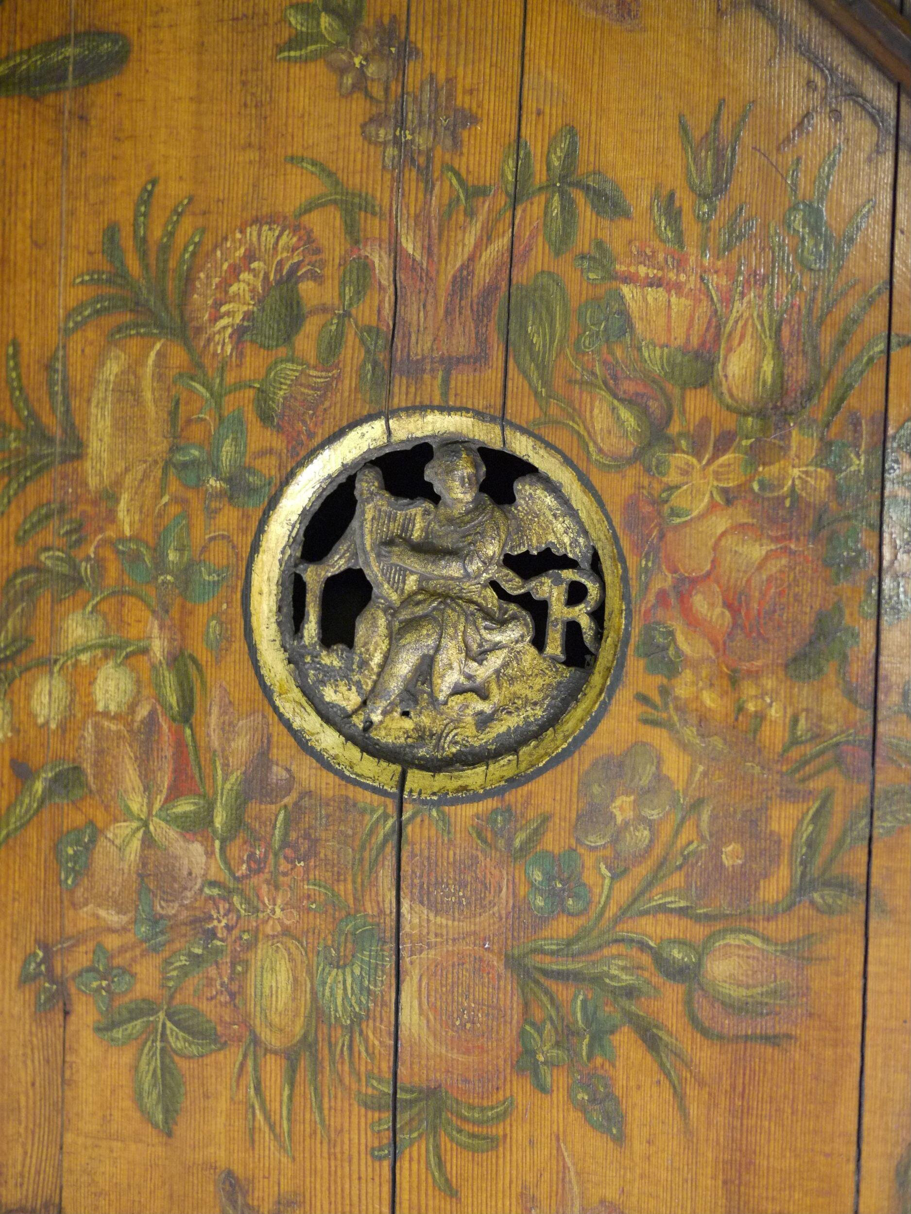 Close-up of a harpsichord soundboard featuring a decorative gilded soundhole carved with a relief of an angel playing a harp. The surrounding wooden surface is richly painted with colorful floral motifs, including tulips, roses, and other blossoms in shades of red, yellow, and green.