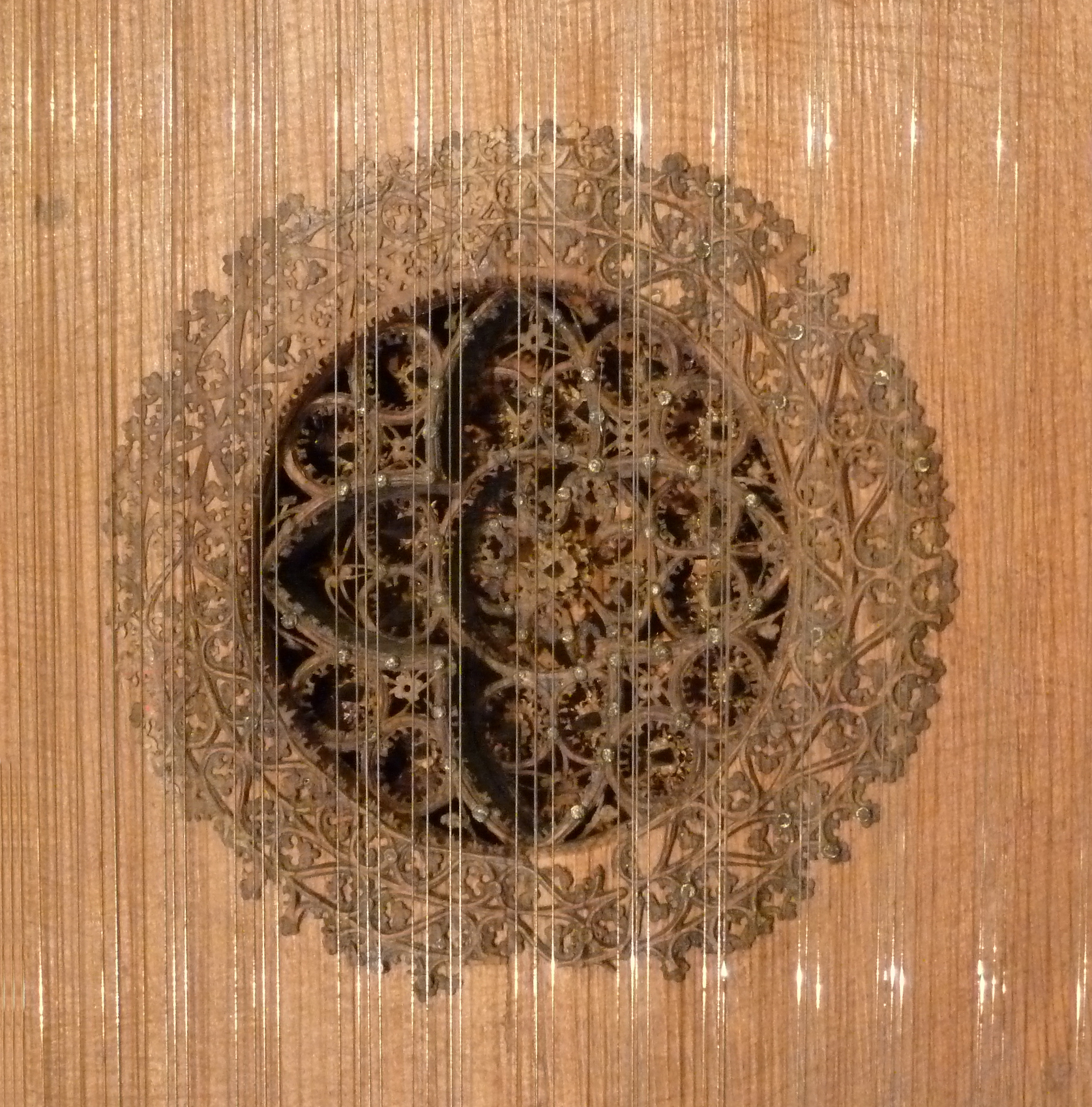 Close-up of an intricately carved wooden rose soundhole on a virginal. The delicate lattice design features interwoven floral and geometric patterns forming concentric circular layers. The strings of the instrument run vertically across the ornate rosette, which is set into a finely grained wooden soundboard.