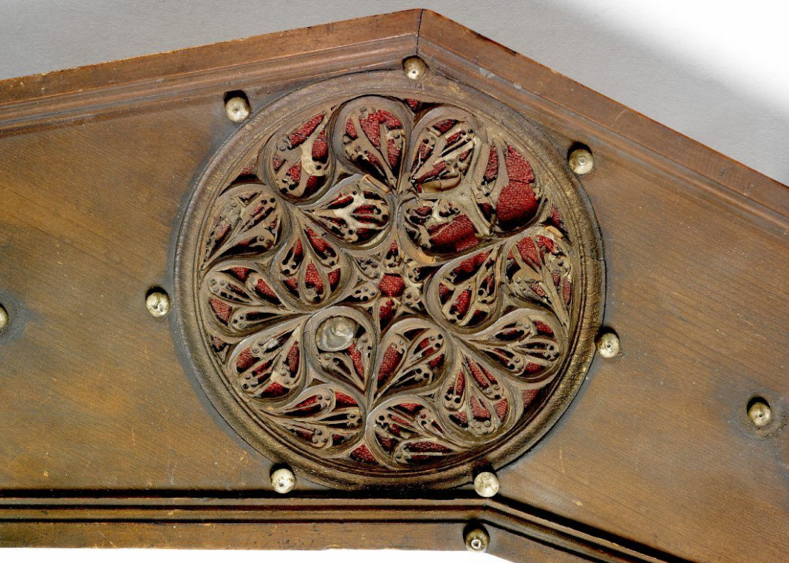 Close-up of an ornate carved wooden rose soundhole on a clavichord. The circular rosette features an intricate interlaced floral and vine motif, set against a red fabric background visible through the openwork design. The wood around the rose is dark and finely finished, with small brass pins securing the decorative panel.