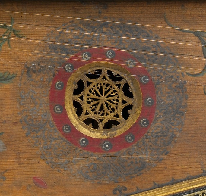 Close-up of a decorative soundhole rose on a Renaissance virginal. The intricate circular carving features a gilded floral and geometric pattern, surrounded by a gold and red painted border with small black-and-white ornamental dots. Delicate blue-gray foliage motifs and thin string lines frame the rosette on the wooden soundboard.