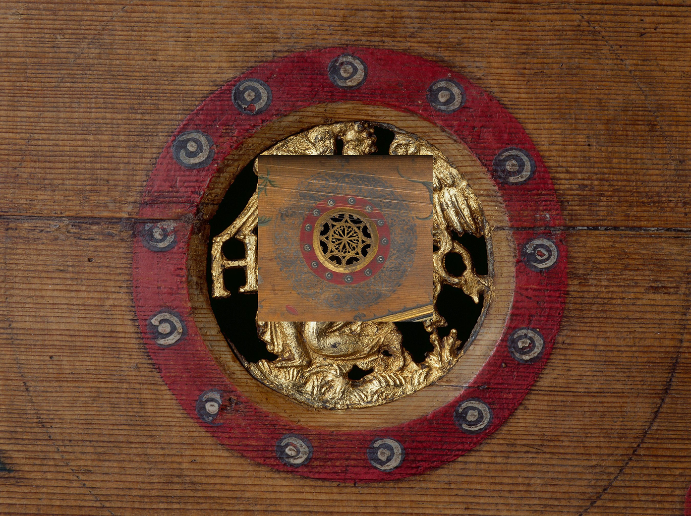 Close-up of an ornate virginal soundhole featuring layered gilded decorations. The outer ring is painted red with alternating black and white circular motifs, framing a gold relief carving that includes an angel and other intricate details. At the center is a smaller inset image showing a decorative rose with a similar design—gold filigree surrounded by a red border and delicate blue-gray foliage patterns—creating a visual echo of the larger soundhole motif.