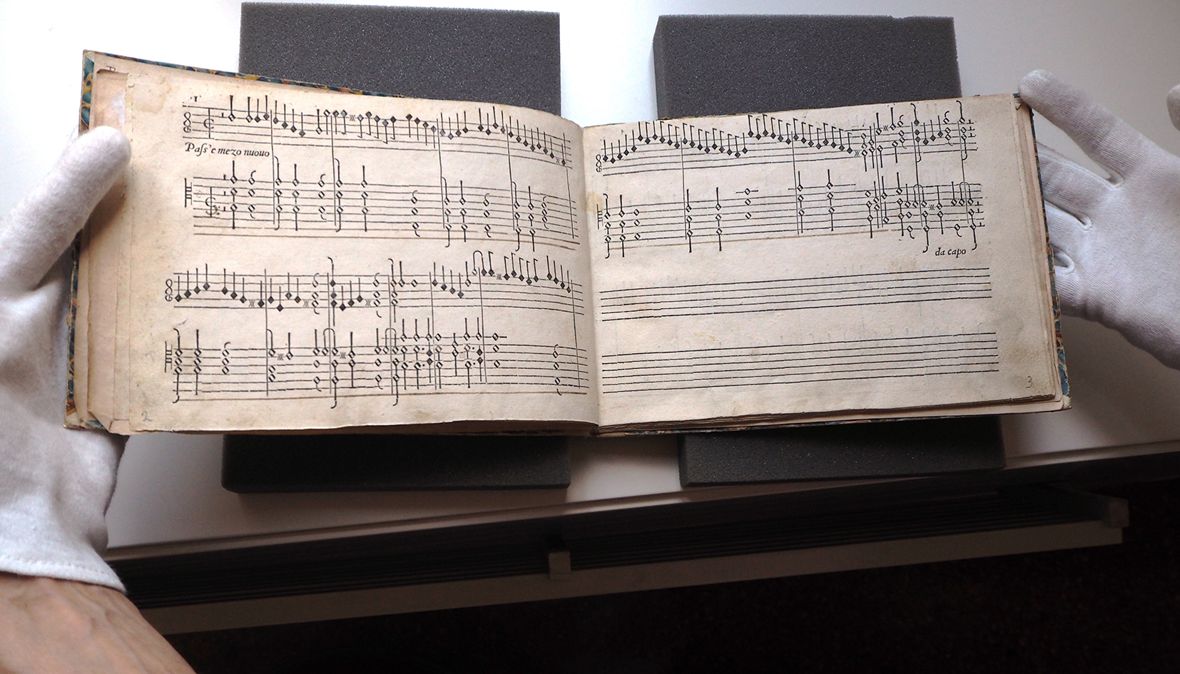 An open 16th-century music book showing two pages of keyboard notation. The staves are filled with dense writing and clear black notes, labelled “Pass’e mezzo nuovo” on the left page. The book is handled with white gloves and supported by foam mounts, indicating archival preservation.
