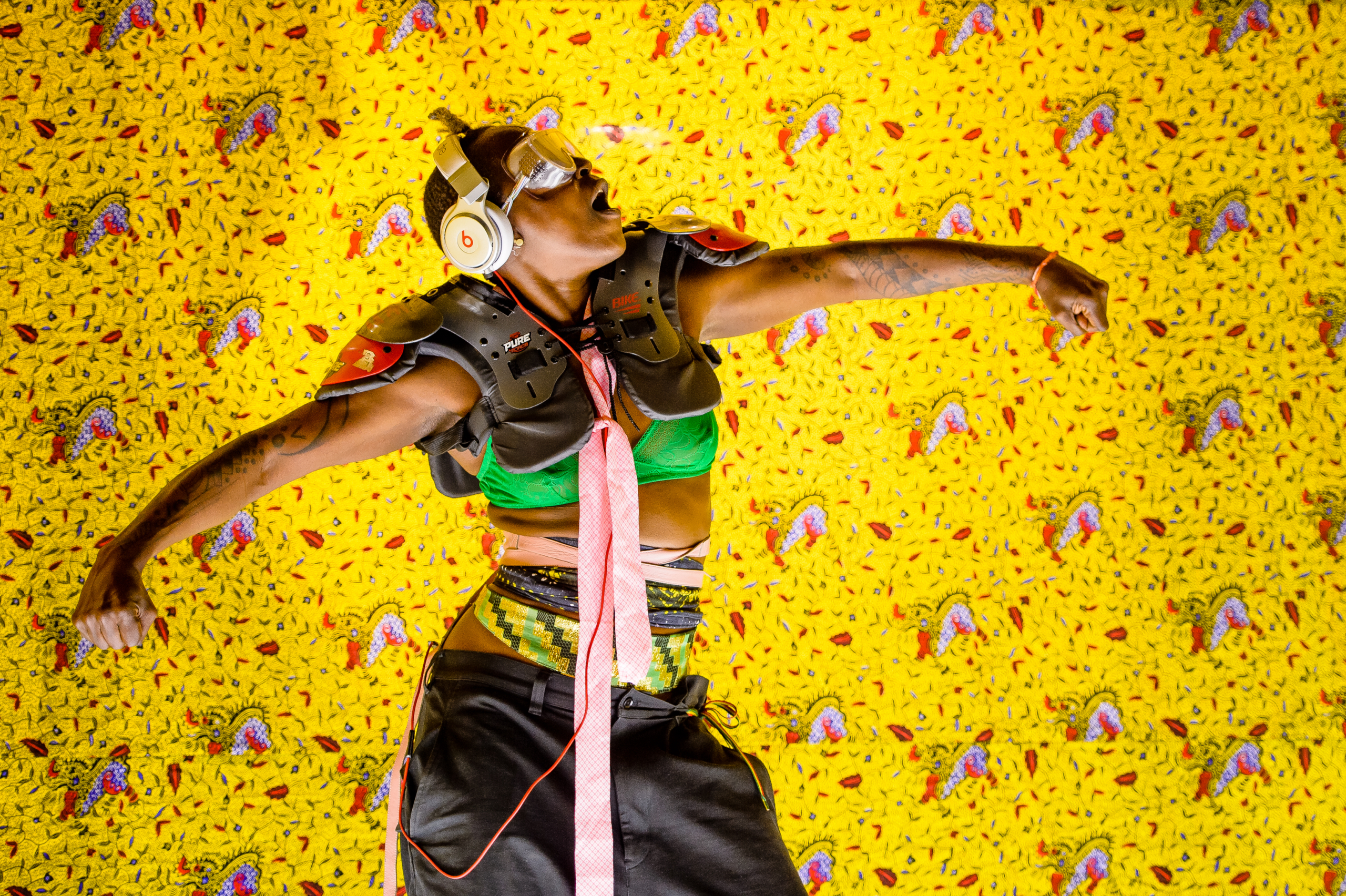 A highly saturated color photograph featuring a performance artist posing energetically against a bright yellow background patterned with abstract red, blue, and black insect-like figures. The performer wears shoulder pads/body armor, a neon green sports bra, black pants worn low, and a patterned athletic belt. They have large, silver goggles pushed up on their head and white headphones over their short hair. The figure has tattoos on both arms and is captured mid-movement with both arms extended and mouth open as if yelling or singing.