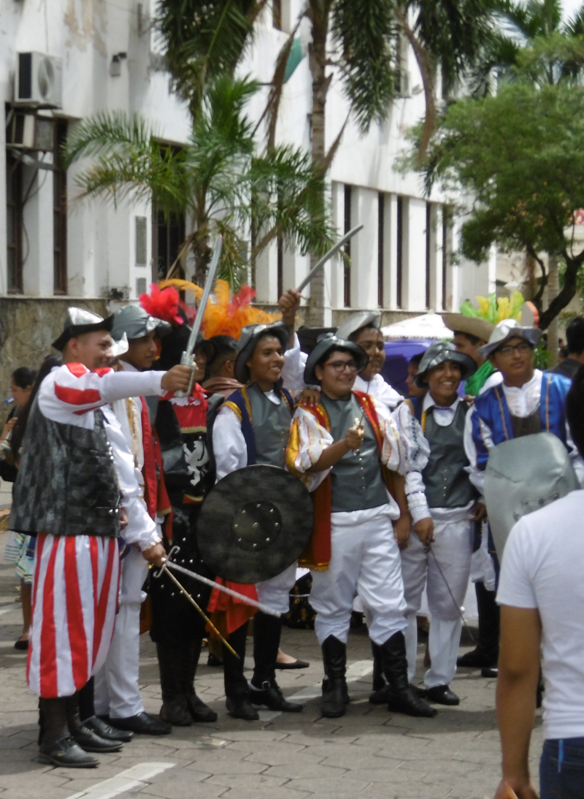 A color photograph showing a group of young men and teenagers participating in a historical or cultural street parade. They are dressed in elaborate, colorful costumes reminiscent of 16th or 17th-century European soldiers, possibly Spanish conquistadors or colonial militia, featuring vests, white breeches, capes (red, blue), tall boots, and helmets/hats with plumes. Many are carrying prop swords and shields. The setting is an urban street in front of white colonial-style buildings with palm trees and a crowd of onlookers, likely in Latin America during a festival or during carnival.