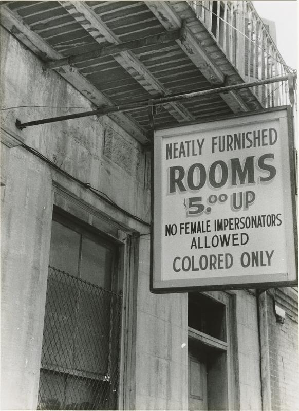 A black and white photograph showing a sign hanging from the overhang of an old building. The sign is rectangular with dark text on a light background reading "NEATLY FURNISHED ROOMS / $5.⁰⁰ UP / NO FEMALE IMPERSONATORS ALLOWED / COLORED ONLY". The building exterior is plain stone and the windows are covered with a wire mesh or grate.