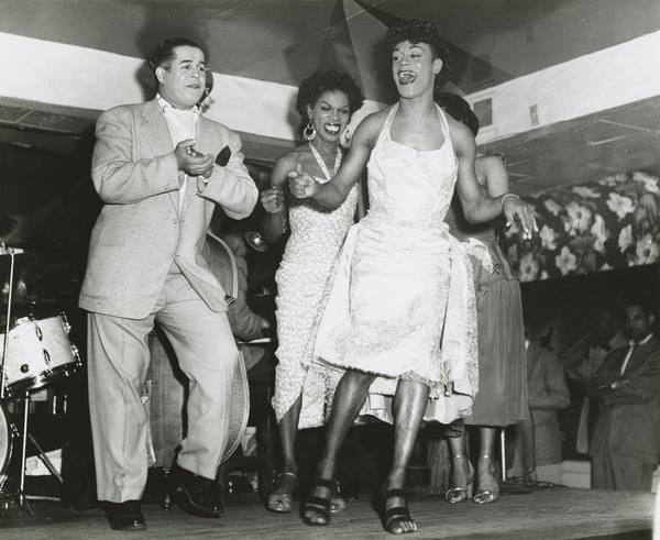 A black and white photograph showing three central figures dancing enthusiastically on a stage. The person on the right wears a light-colored suit, the person in the middle wears a patterned dress with hoop earrings and the person on the right, in front of the others, wearing a light-colored halter dress and high heels. The background features floral-patterned curtains and a partial view of a drum set on the far left, suggesting a lively nightclub or cabaret performance.