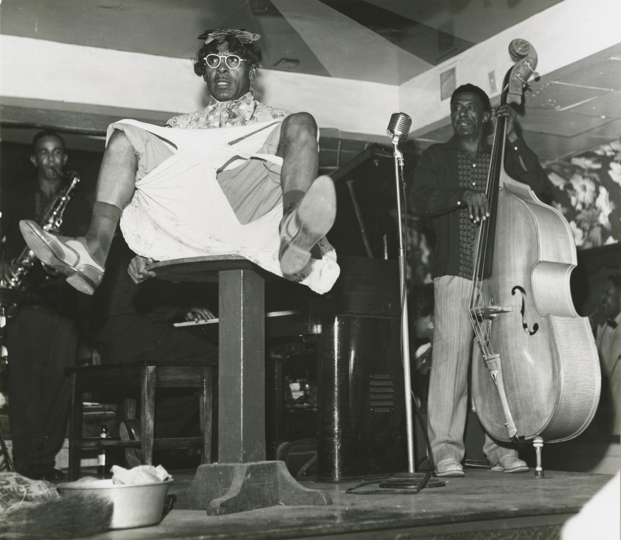 A black and white photograph of three musicians performing on a stage. In the foreground, a person is seated atop a tall, square table with their legs spread in the air, revealing the soles of the shoes and light-colored undergarments beneath a patterned shirt. They wear a small hat and round glasses. To the left, a man plays the saxophone. To the right, a man plays a double bass (upright bass), standing near a microphone. The background is dimly lit and suggests a club setting.