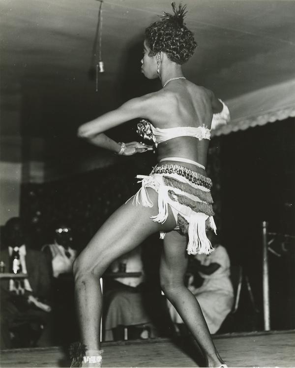 A black and white photograph of a dancer performing on a stage, viewed from the rear/side. The dancer is wearing a minimal two-piece costume, including a fringed belt/skirt around and a brassiere-style top. The dancer is mid-movement, leaning forward with their legs bent. In the dimly lit background, several persons are seated at tables watching the performance.
