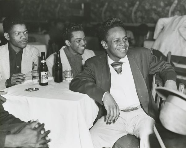 A black and white photograph of three people seated around a small, white-linened table in what appears to be a club or restaurant. The person on the right, the main focus, smiles widely, leaning back in the chair while wearing a dark jacket, light trousers, a light shirt, and a striped tie. The two men behind the table also wear jackets. The table holds two beer bottles and glasses.