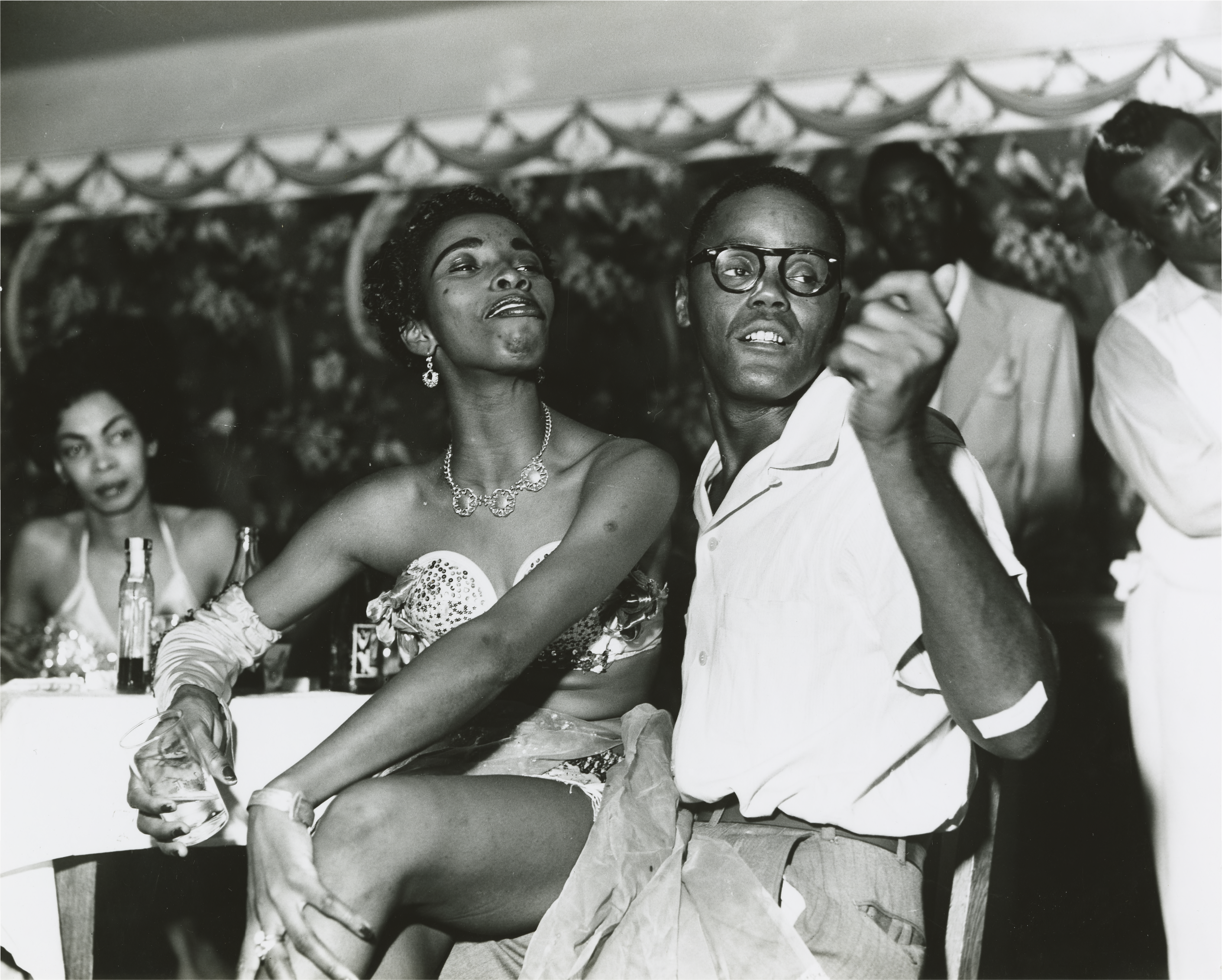A black and white photograph of two people seated closely together at a small table in a nightclub setting. The person on the left wears a strapless, sequined top, a layered skirt, one long glove and jewelry, looking up and to the left. The person on the right, wearing a light-colored, short-sleeved shirt and glasses, gestures with their hand while looking in the same direction. In the background, other people are visible, and decorative scalloped fabric hangs above.