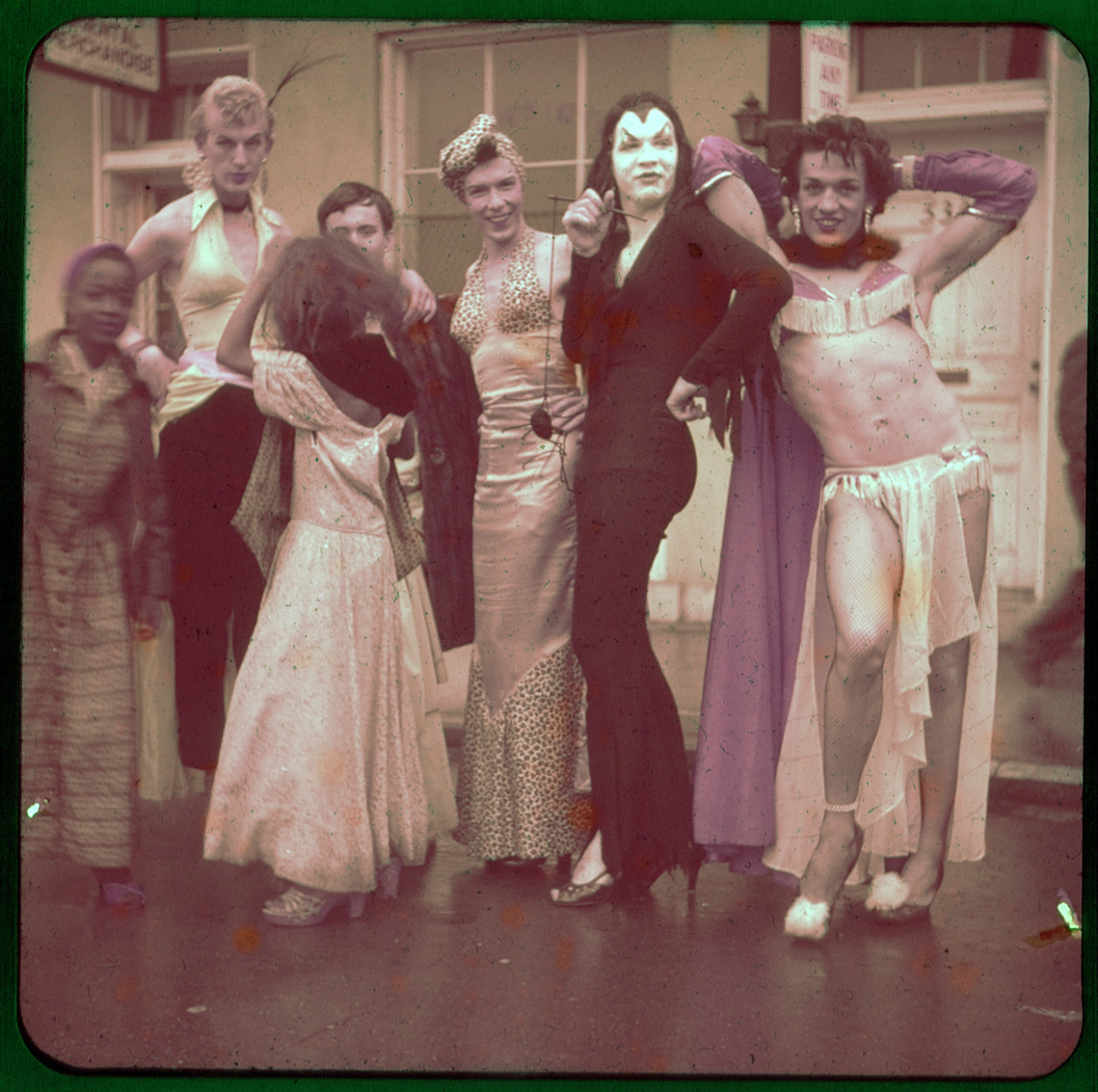 A vintage color photograph of a group of eight people in drag or elaborate costumes, posing closely together outdoors on a wet street. Costumes include: a person in a light top and dark pants; a person in a leopard-print dress; a person dressed in a tight black dress and pale makeup; and a person in a skimpy fringed two-piece costume, fishnet stockings, and heels. On the far left, a young child in a long dress stands next to the group. The overall image has a slight green tint.