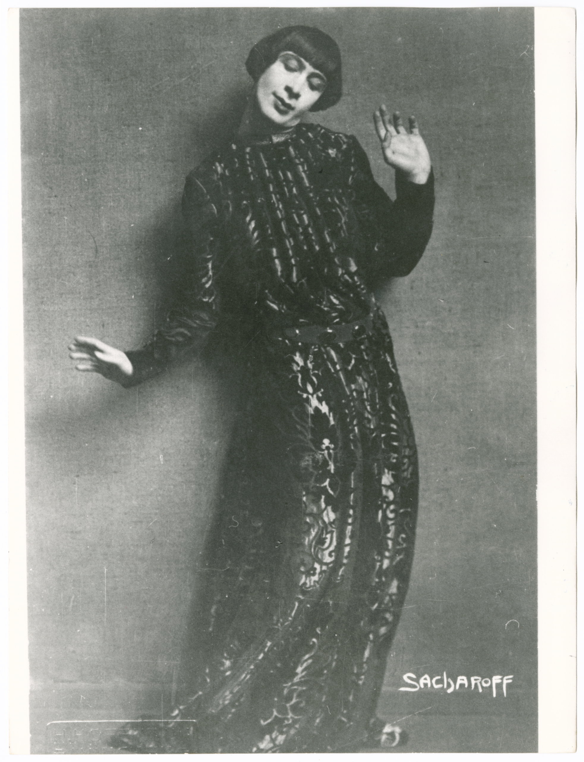 A black and white studio portrait of the dancer Alexander Sacharoff (often billed as 'Sacharoff' on photographs). The figure is dressed in a long, flowing, dark gown with an elaborate patterned or sequined texture and a fashionable bob haircut. Sacharoff is posing dramatically, extending their hands gracefully in a dance movement, with one hand raised near the head and the other lowered. The signature 'SACHAROFF' is printed in the lower right corner. The photo is reflective of early 20th-century avant-garde and aesthetic dance.