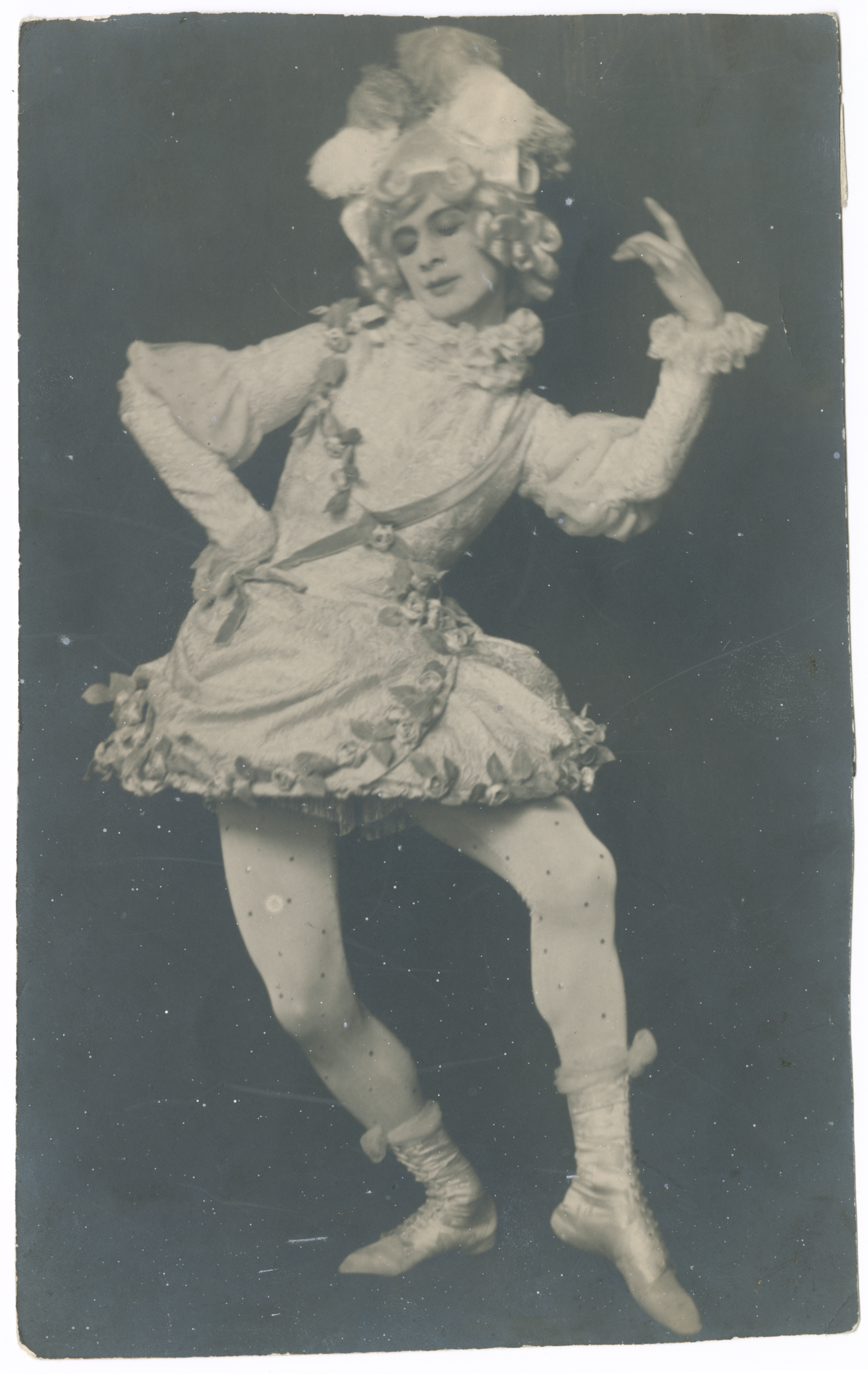 A vintage, slightly faded black and white portrait of a performer in an elaborate theatrical costume. The costume is pale and ruffled, featuring an embroidered or patterned short jacket/tunic with puffed sleeves, frills around the neck and cuffs, and rosettes. The performer wears light-colored tights/stockings (with visible spotting on the photo) and soft ballet slippers with bows, topped by a curly wig and a large plumed hat. The figure is captured mid-pose, leaning back dramatically with one hand raised, embodying a character from a ballet, pantomime, or historical piece, possibly a Baroque-style court figure or jester.