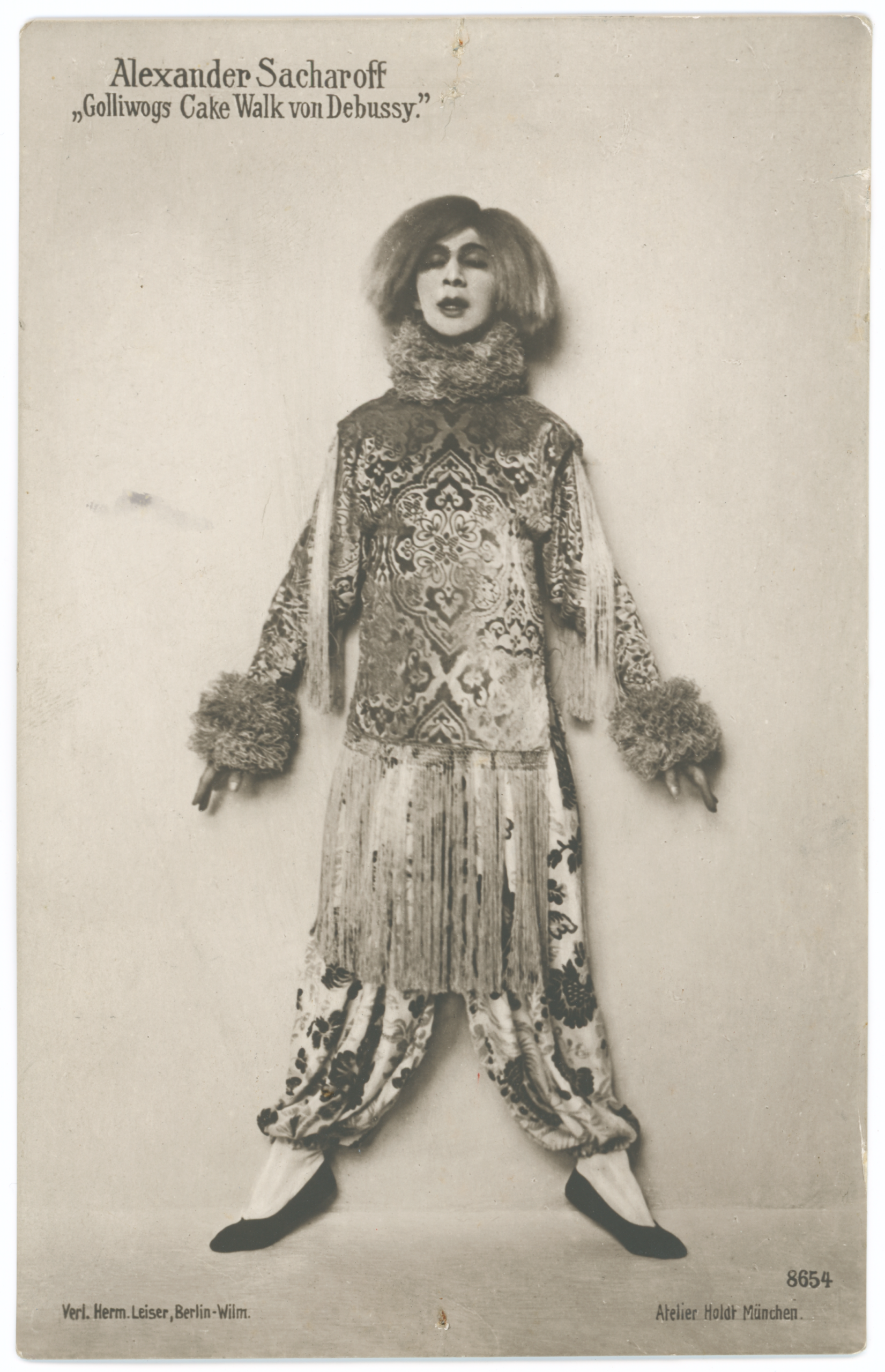 A vintage black and white full-length studio photograph of the dancer Alexander Sacharoff, wearing an elaborate costume for his performance of "Golliwoggs Cake Walk von Debussy." The costume features a voluminous top and harem-style pants made of a richly patterned, possibly brocade or velvet, fabric. It includes a high, ruffled collar, fringe around the waist, and large, fuzzy cuffs at the wrists. Sacharoff is wearing pale makeup and a short, dark, voluminous wig. He stands in a wide, stylized dance pose, with his arms extended and his feet turned out. Text above the image identifies him as "Alexander Sacharoff" and the piece as "»Golliwoggs Cake Walk von Debussy.«" Additional text identifies the publisher and the photo studio.