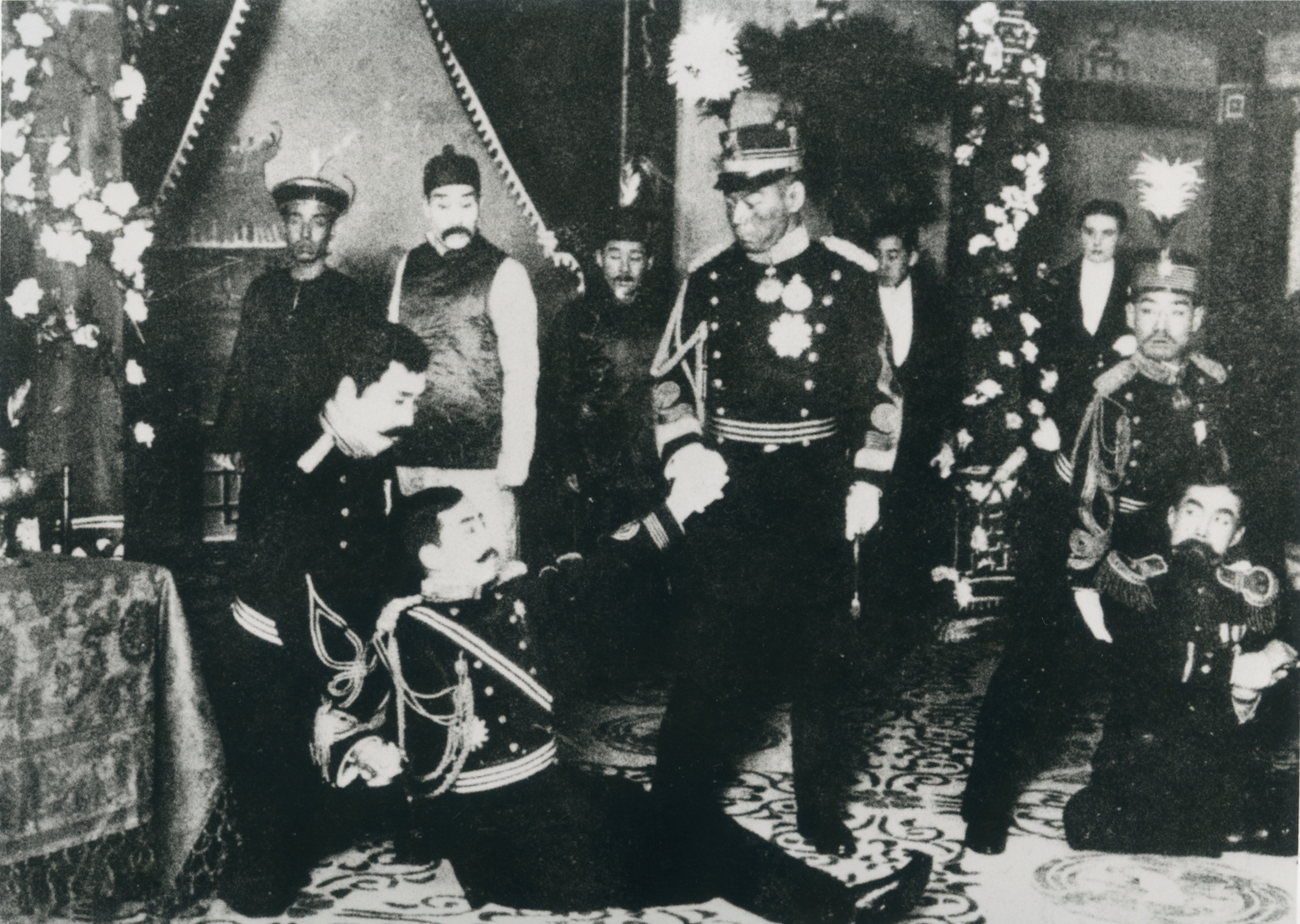 A black and white historical theater photograph taken indoors, showing a formal and solemn ceremony with several men in military and official attire. In the center, actor in a highly decorated military uniform with many medals stands tall, firmly grasping the arm of another actor in a similar, dark military uniform who is kneeling before him. The kneeling actor appears distressed or subservient, with head bowed. Several others, some in military dress and some in traditional formal wear (including hats with turbans or caps), stand solemnly in the background. The room is decorated with fabric drapes and floral arrangements, suggesting a significant political or official event, possibly an apology, surrender, or formal oath.