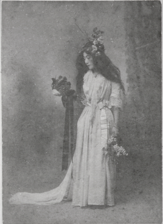 A sepia-toned or heavily faded black and white portrait, typical of the late 19th or early 20th century, showing someone in a long, flowing, light-colored feminine dress (possibly white or cream), standing in a pensive or theatrical pose. The dress features a high waistline and ribbon accents. The figure has very long, dark hair flowing over her shoulders, and flowers woven into their hair. They hold two small bouquets of flowers and long ribbons drape from their hands. The image has a soft focus and is set against a plain studio background, suggesting an aesthetic or theatrical costume, possibly for a classical or mythological role.