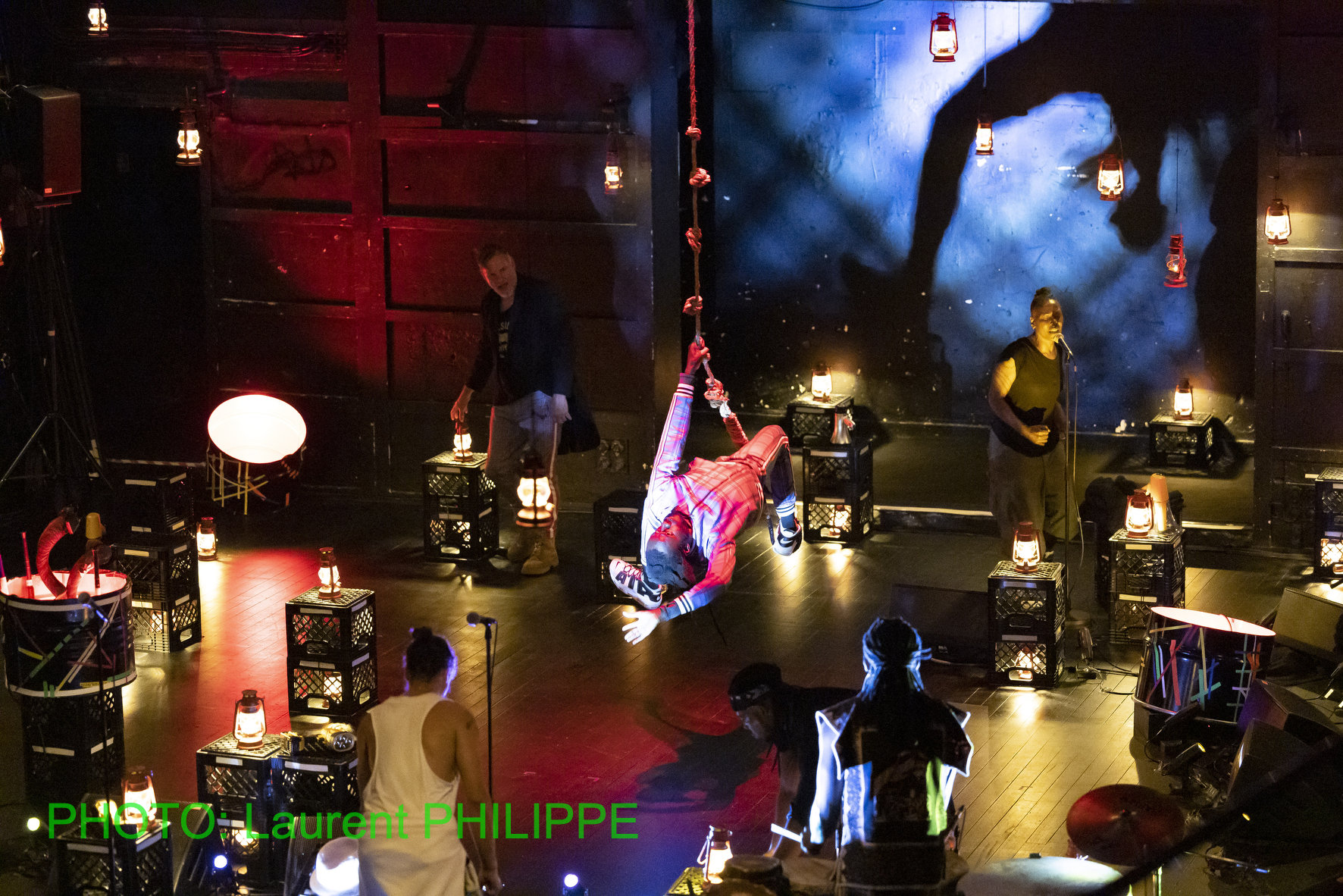 A dark color photograph capturing a stage performance heavily decorated with milk crates, lanterns, and hanging ropes. In the center, a performer in a plaid shirt and dark pants is suspended mid-air, hanging upside down from a thick rope. Two other standing performers are visible: one on the left holding a lantern, and one on the right singing into a microphone. The stage is lit with deep red and blue spotlights, casting strong shadows and highlights on the polished floor. Several small, glowing lanterns hang from above and rest on the scattered milk crates, which function as set pieces, risers, and platforms for instruments/props. A figure in a white tank top is visible in the foreground with their back to the camera. The name "PHOTO: LAURENT PHILIPPE" is overlaid on the lower left.