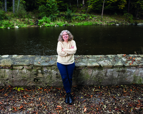 Die Komponistin Astrid Spitznagel lehnt an einer niedrigen Steinmauer. Im Hintergrund fließt ein Fluss. Das gegenüberliegende Ufer ist von einem Wald gesäumt.