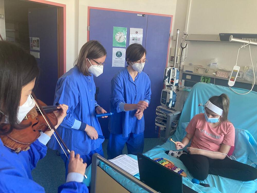 Drei in blauen Krankenhausgewändern gekleidete Personen musizieren (Geige, Claves) mit einem im Krankenbett sitzenden Mädchen das auf einem Xylophon spielt.   
