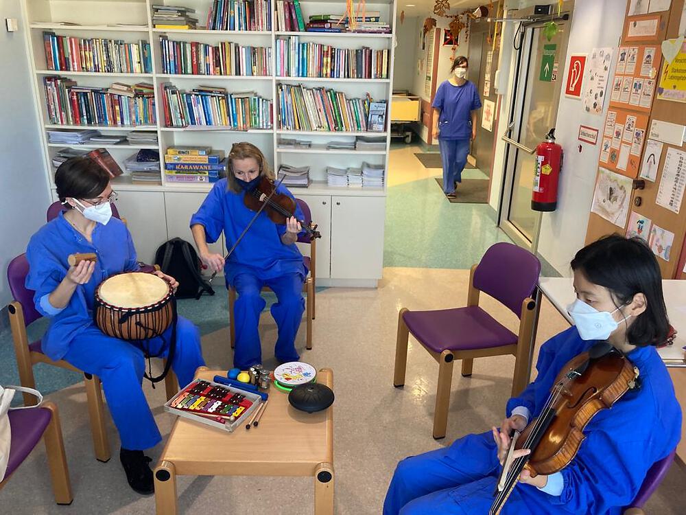 Foto von drei in blauen Krankenhausgewändern gekleideten Personen die  gemeinsam im Krankenhaus musizieren (Geige, Claves, Trommel).