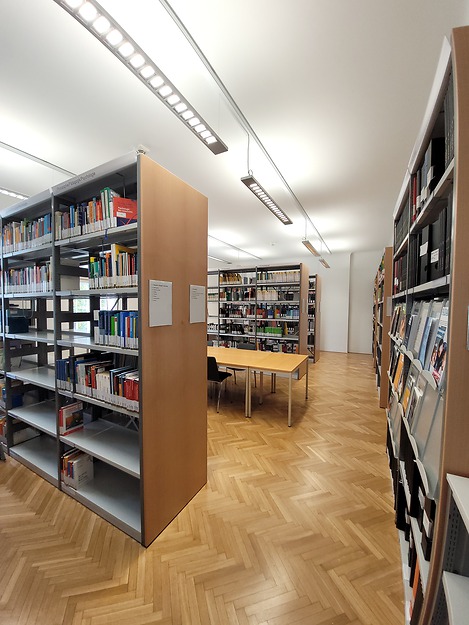 Foto der Bibliothek mit mehreren Regalen freistehend sowie an der Wand, Gängen dazwischen und einem Tisch mit Sessel 
