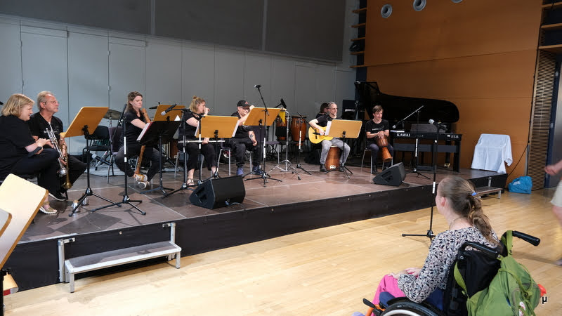 Foto von acht Musikern auf der der Bühne, die gerade nicht spielen. Im Vordergrund ist eine Zuhörerin im Rollstuhl. 