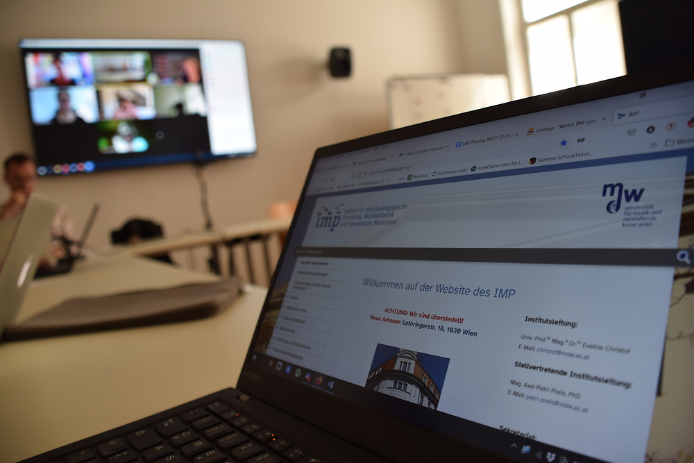 Foto eines Laptops auf dem die Homepage-Startseite des Institus zu sehen ist, im Hintergrund sieht man die online Teilnehmer am Plenum. 