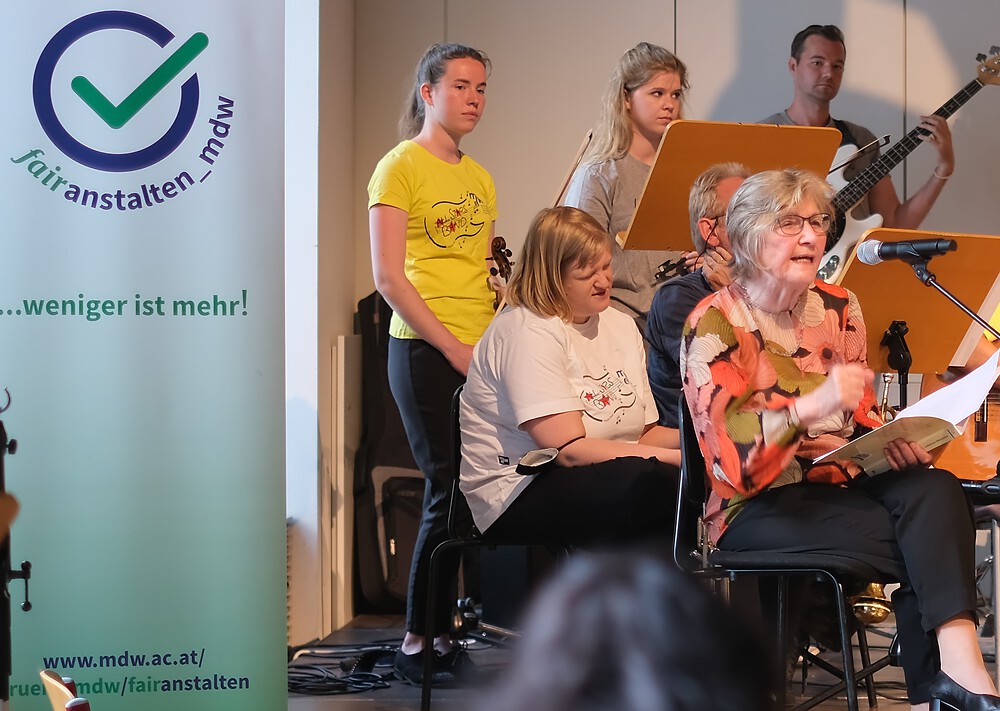 Foto von der Lesung von Helga mit Passagen aus dem Buch „Inklusiv musizieren – Praxis, Pädagogik, Ästhetik (am Beispiel der All Stars Inclusive Band Wien)“. Helga sitzt im Vordergrund, hinter und neben ihr sind Musiker. Am linken Bildrand sieht man das FairAnstaltungslogo. 