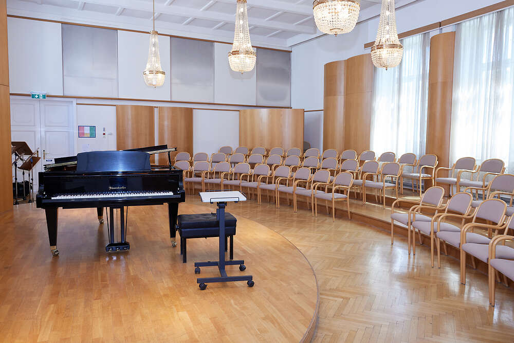 Foto.  Franz Liszt-Saal. Konzertsaal mit Klavierflügel auf erhöhtem Podium und im Halbkreis angeordneten Sesseln für das Publikum. Beleuchtung u.a. Kristallluster. 