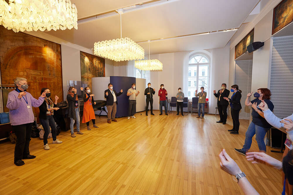 Teilnehmer_innen am Symposium die einem kleineren Saal im Kreis stehen und ihre Hände in die Luft halten. 