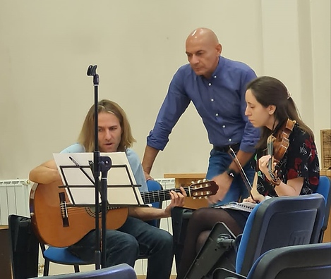 Das Foto zeigt drei Personen, Bernhard sitz mit seiner Gitarre hinter einem Notenständer, daneben sitz eine Frau mit einer Geige und dahinter steht ein Mann. Alle drei sehen auf die am Notenpult befindlichen Noten. 