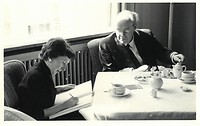 Sie sehen eine Schwarz-Weiß-Fotografie, auf der Herta Blaukopf an einem Tisch mit Arnolt Bronner sitzt und in ein Heft schreibt.