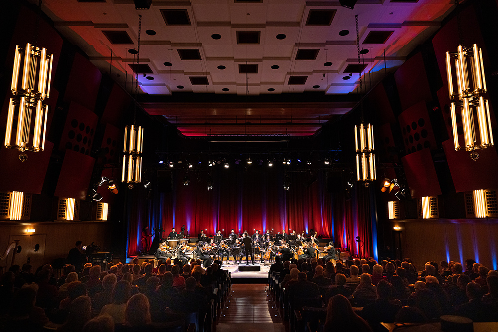 Foto vom letztjährigen Konzert im bunt beleuchteten, vollbesetzten Sendesaal des Radiokulturhauses. Ein Studierender im Anzug dirigiert auf der Bühne das Orchester.